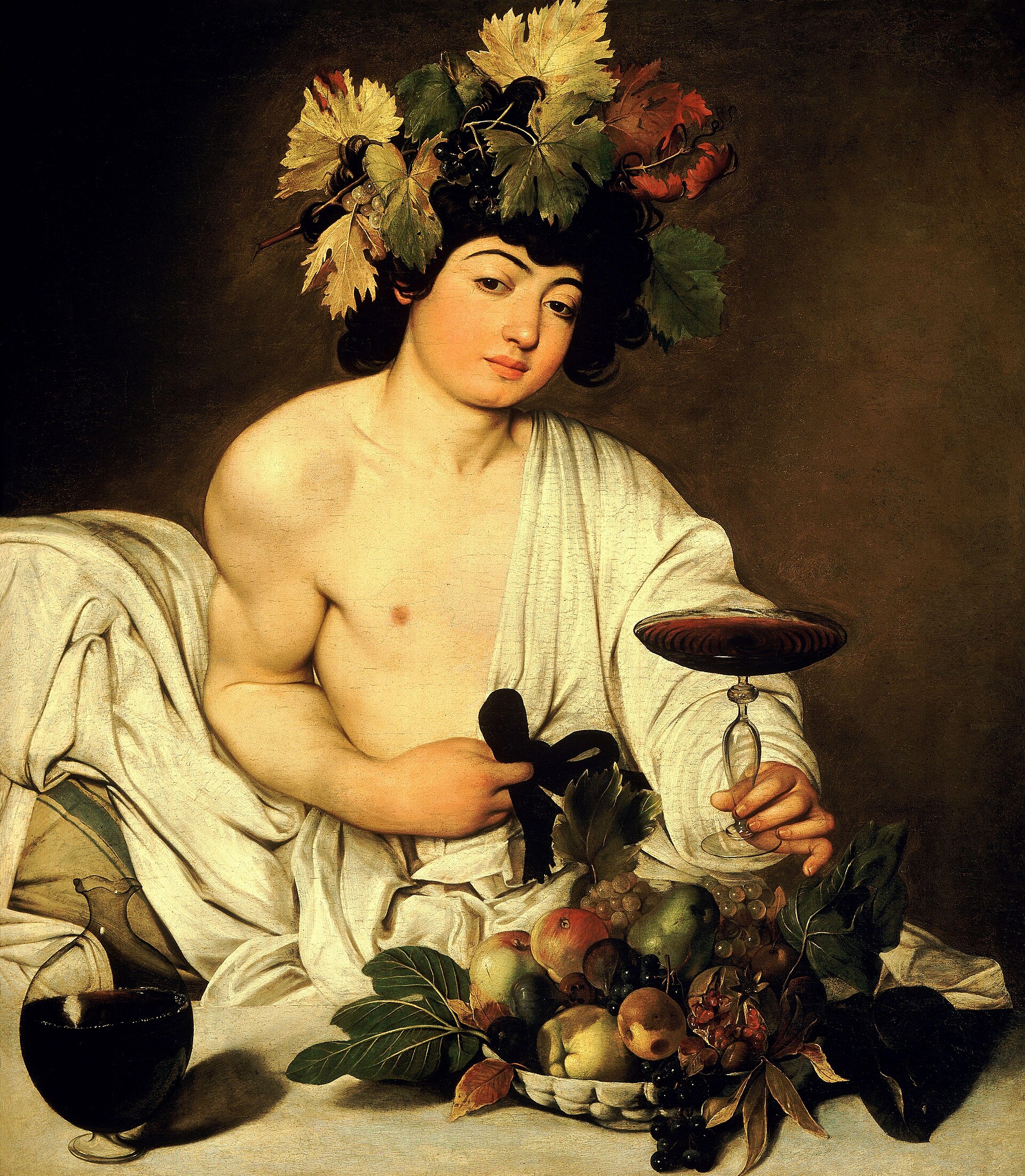 A youthful Bacchus reclining behind a stone table, bare-shouldered in a loose white robe and crowned with an autumnal wreath of grapevine leaves and dark grape clusters. He holds out a shallow footed glass of red wine in his left hand while the fingers of his right toy with the black bow of his sash. A large glass carafe of red wine stands at the lower left, and a heaped bowl of fruit — grapes, apples, a pear, a split pomegranate and figs — sits before him against a dark ground.