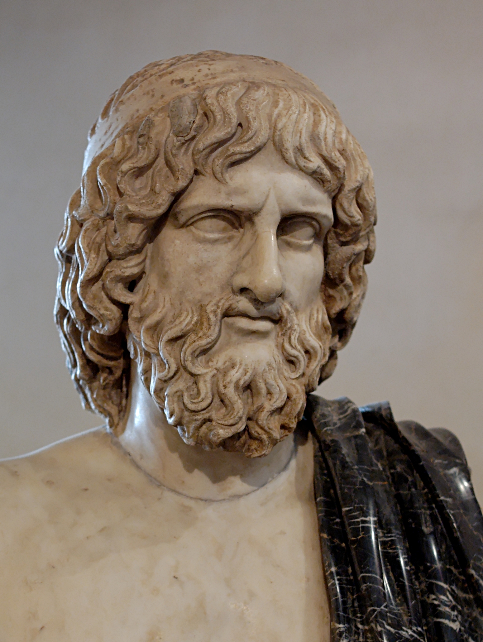 A marble bust of Hades — a mature, heavy-bearded god with thick wavy hair falling to the shoulders, deep-set blank eyes and a furrowed, stern brow, gazing off to the side. A dark near-black marble mantle (a modern restoration) is draped over his right shoulder, set against a plain grey ground.