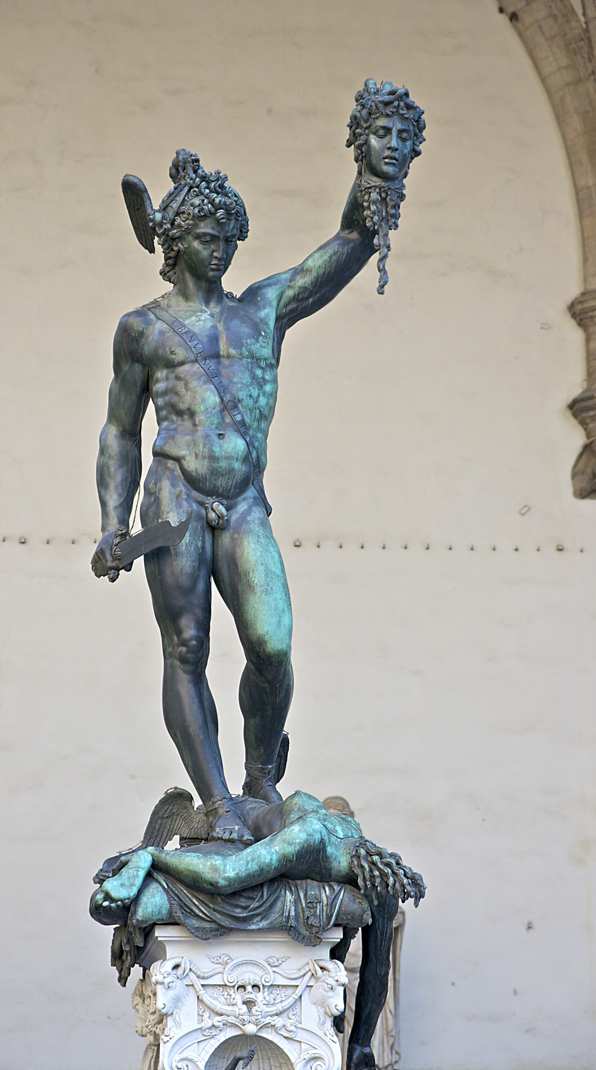 A nude young hero in a winged cap holds aloft the severed, snake-haired head of Medusa in his raised left hand and a curved sickle in his lowered right, his own gaze cast down and away from the trophy; the Gorgon's decapitated body slumps beneath his feet on an ornate pedestal against a plain pale wall — Benvenuto Cellini's bronze Perseus with the Head of Medusa in the Loggia dei Lanzi, Florence.