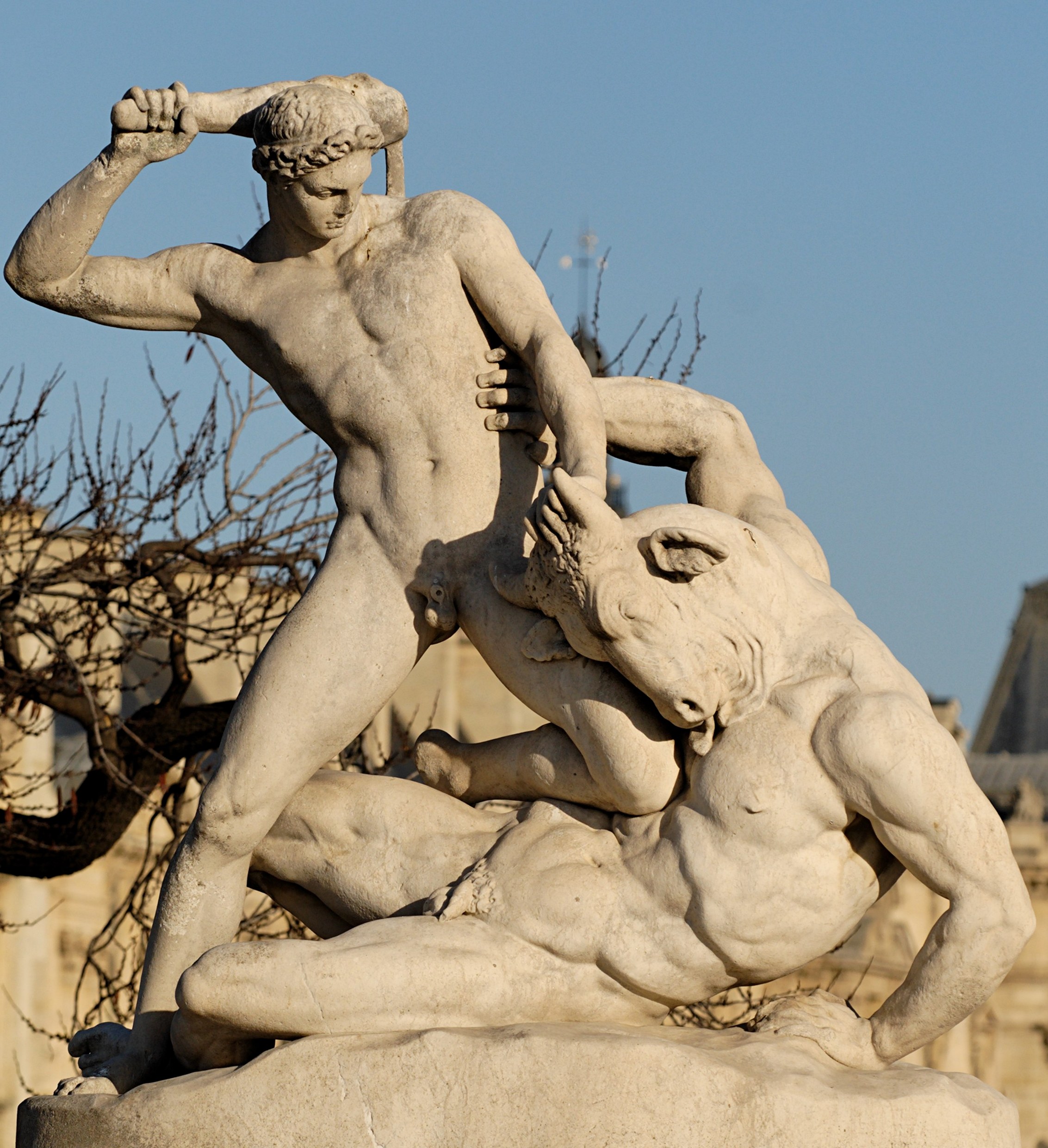 A white marble statue of a nude, laurel-crowned Theseus lunging forward with a club raised overhead in one hand, gripping the horned head of the bull-headed Minotaur with the other as the beast collapses beneath him over a fallen youth, set against a soft-focus ground of bare trees and pale stonework.