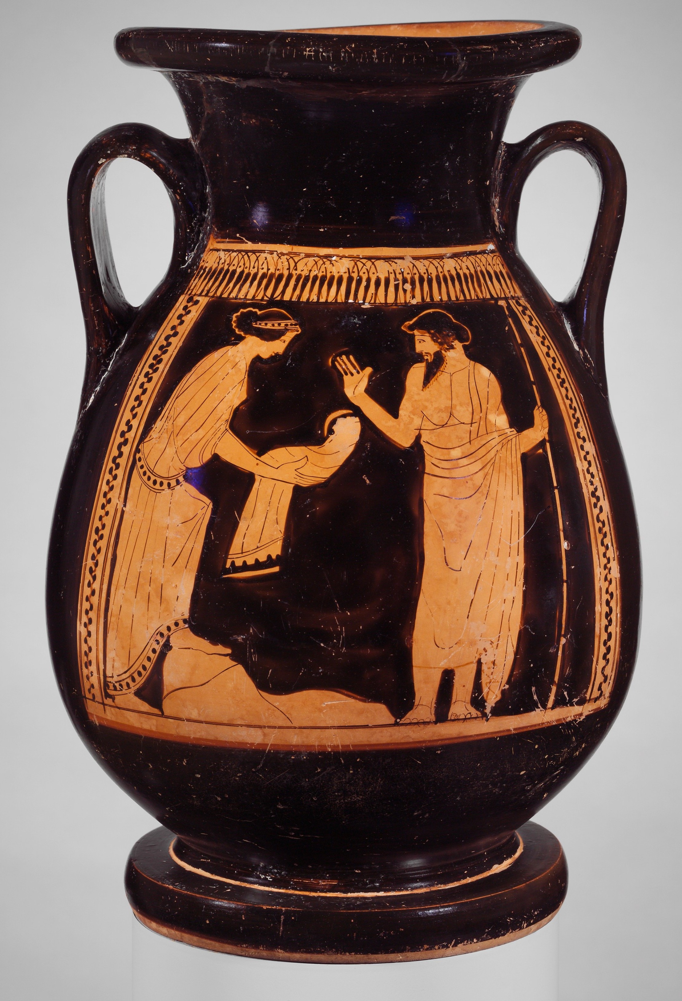 An Attic red-figure pelike, the whole black-glazed vessel standing against a plain grey ground, its belly panel bordered by a band of tongues above and running dot-chains at the sides. In the scene the Titaness Rhea, at left in a long pleated chiton and mantle with her hair bound in a fillet, steps forward and holds out in both hands the stone wrapped in swaddling clothes; facing her at right the bearded Kronos, in a himation that leaves one shoulder bare and a soft cap, grips a long sceptre in his trailing hand and raises his other hand to receive the bundle he believes is his newborn son — the swaddled-stone deception that founds the ruling family.